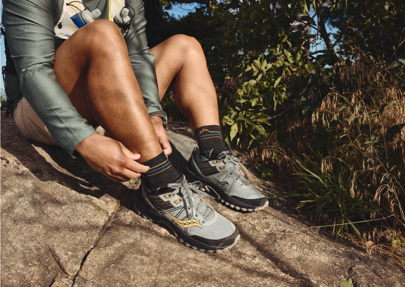 Trail runner adjusting Swiftwick socks while sitting on a rock outdoors.