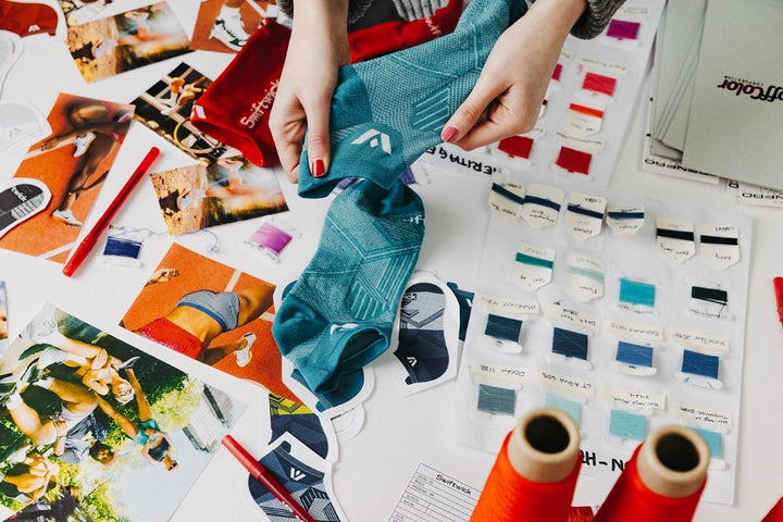Overhead view of a design table with yarns, product photos, pens, and hands stretching a sock.