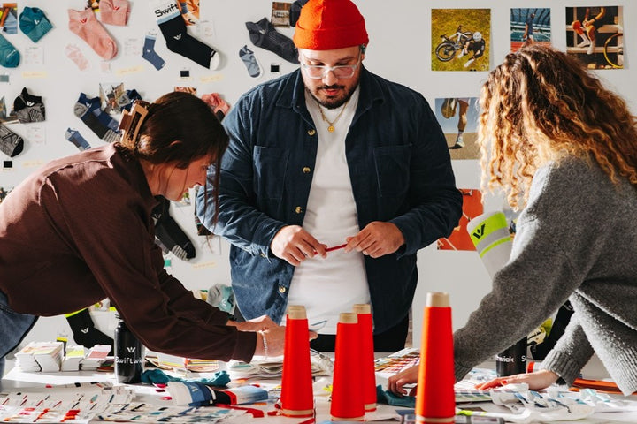 Three people working at a design table covered in yarns, socks, and color swatches with a white wall full of pinned socks and photos in the background.