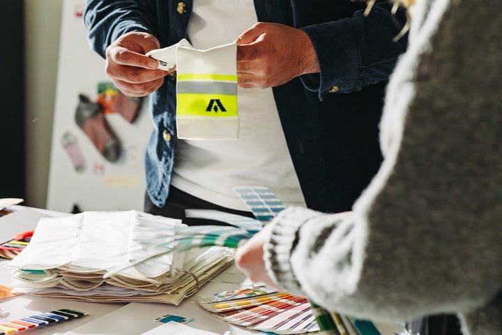 Closeup of a person holding up a sock while another, out of focus, holds color swatches with a table of swatches below.