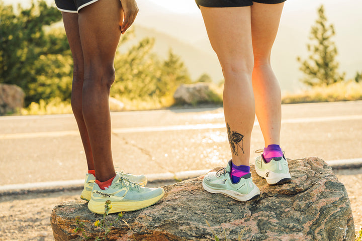 Closeup of two runners in Swiftwick socks and running shoes, with a road and sunset in the background.