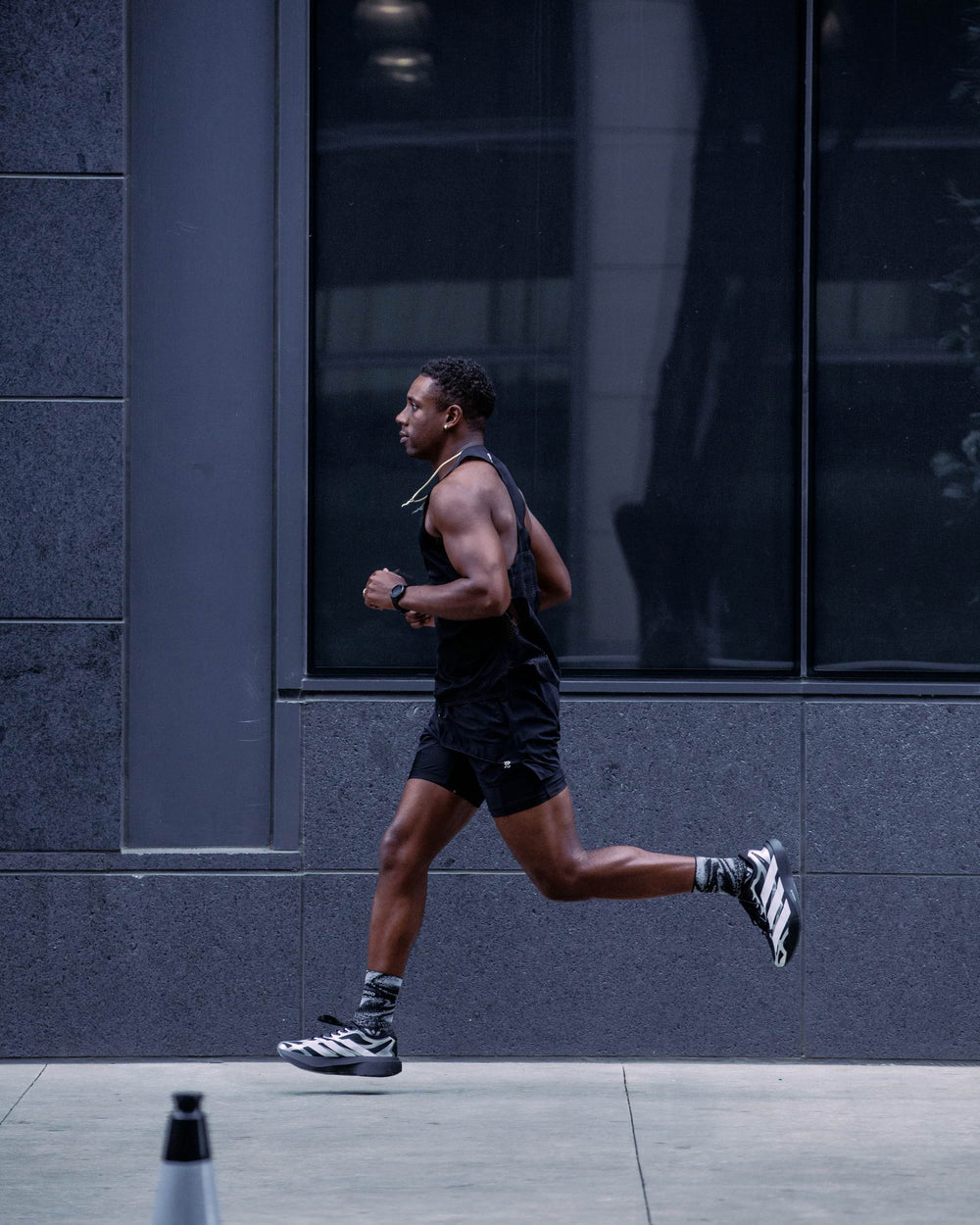 A man running in a black athletic tank top and shorts with black and white running shoes, captured in motion against a dark gray modern building facade.