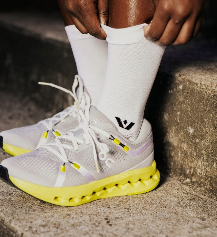 Person wearing white sneakers with yellow soles and a brand logo, sitting on a concrete surface.