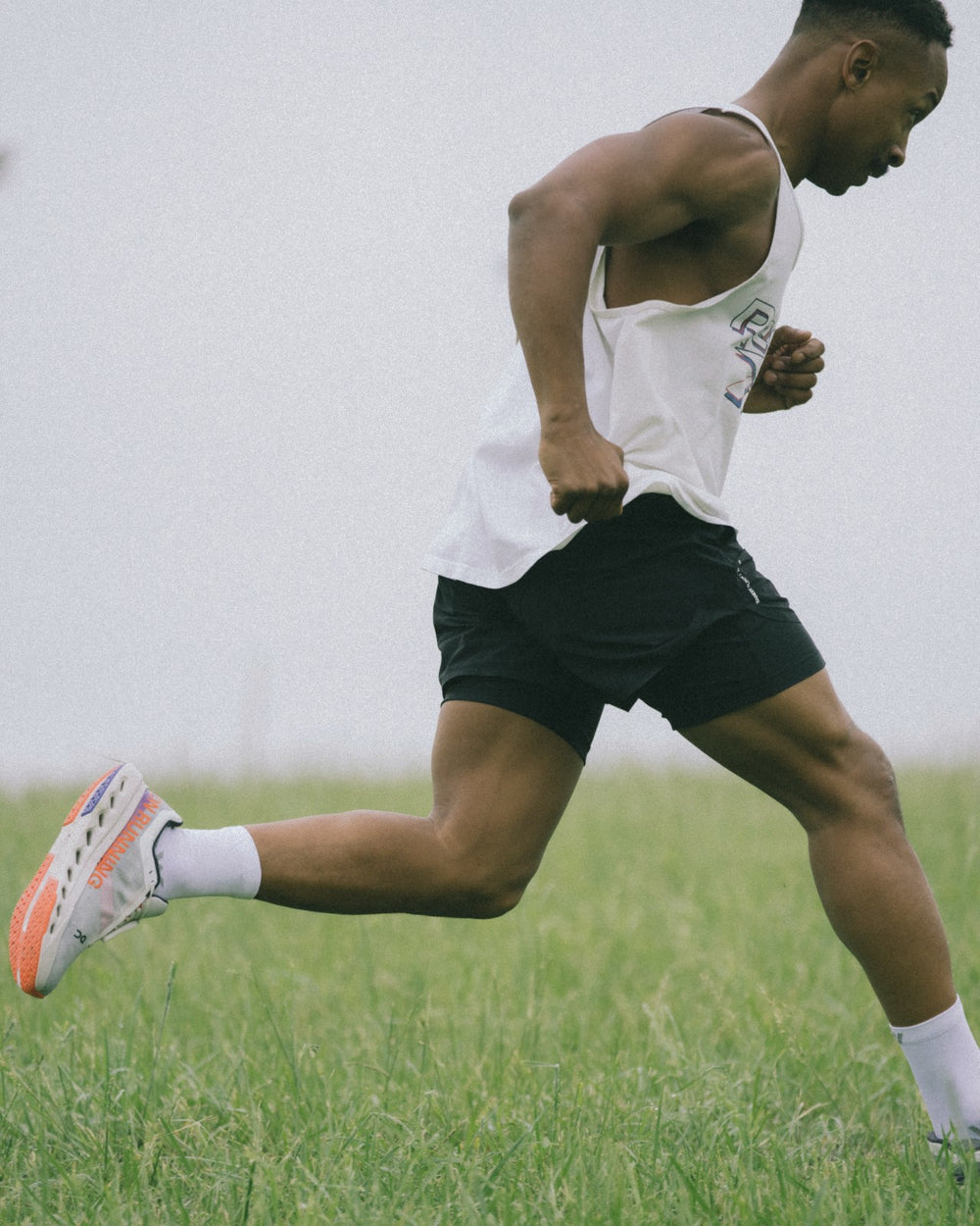 A man running through a grassy field in a white tank and black shorts, wearing white Swiftwick socks and running shoes.