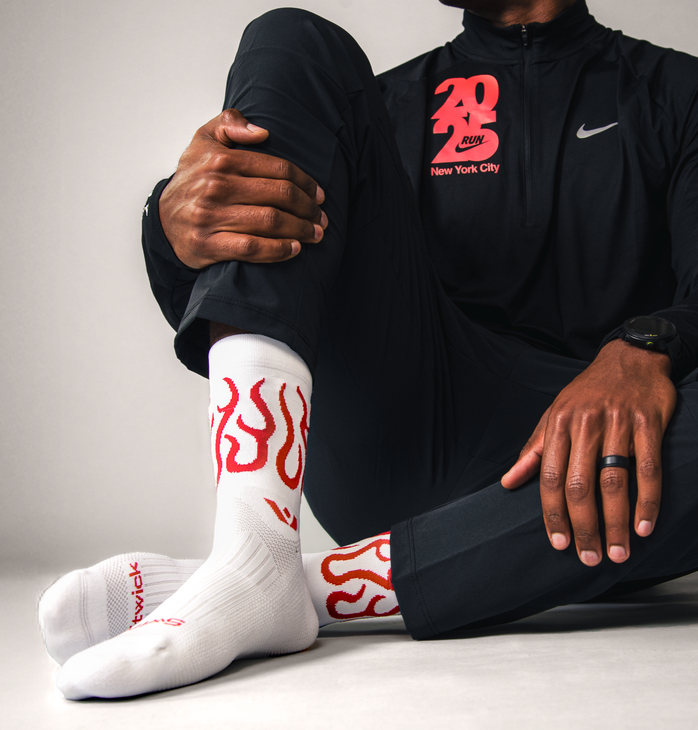 Person sitting down on white background wearing a black Nike tracksuit and Swiftwick Vision Mid-Crew Flames socks.